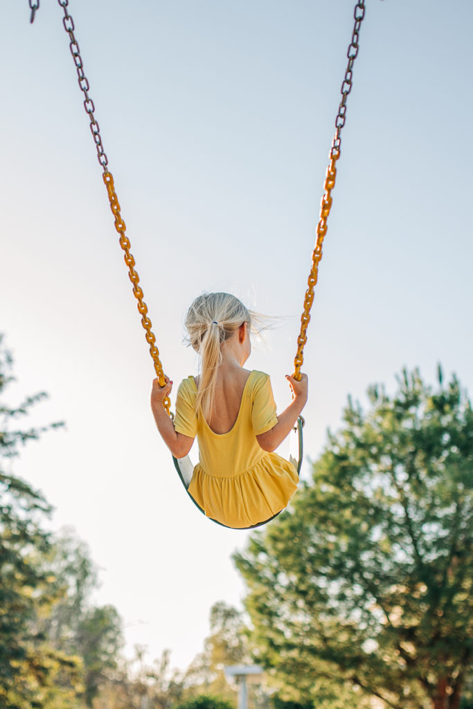 Family Photographer, a young girl in a yellow dress swings on the swings, trees and sky before her