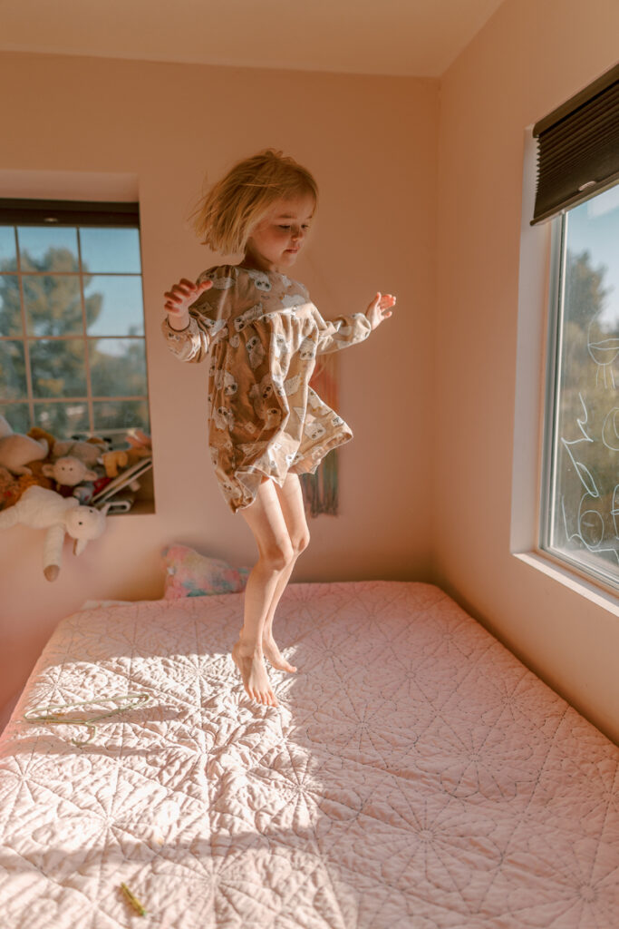 Family Photographer, a young girls jumps up down in the bed, sunlight from a window shines on her