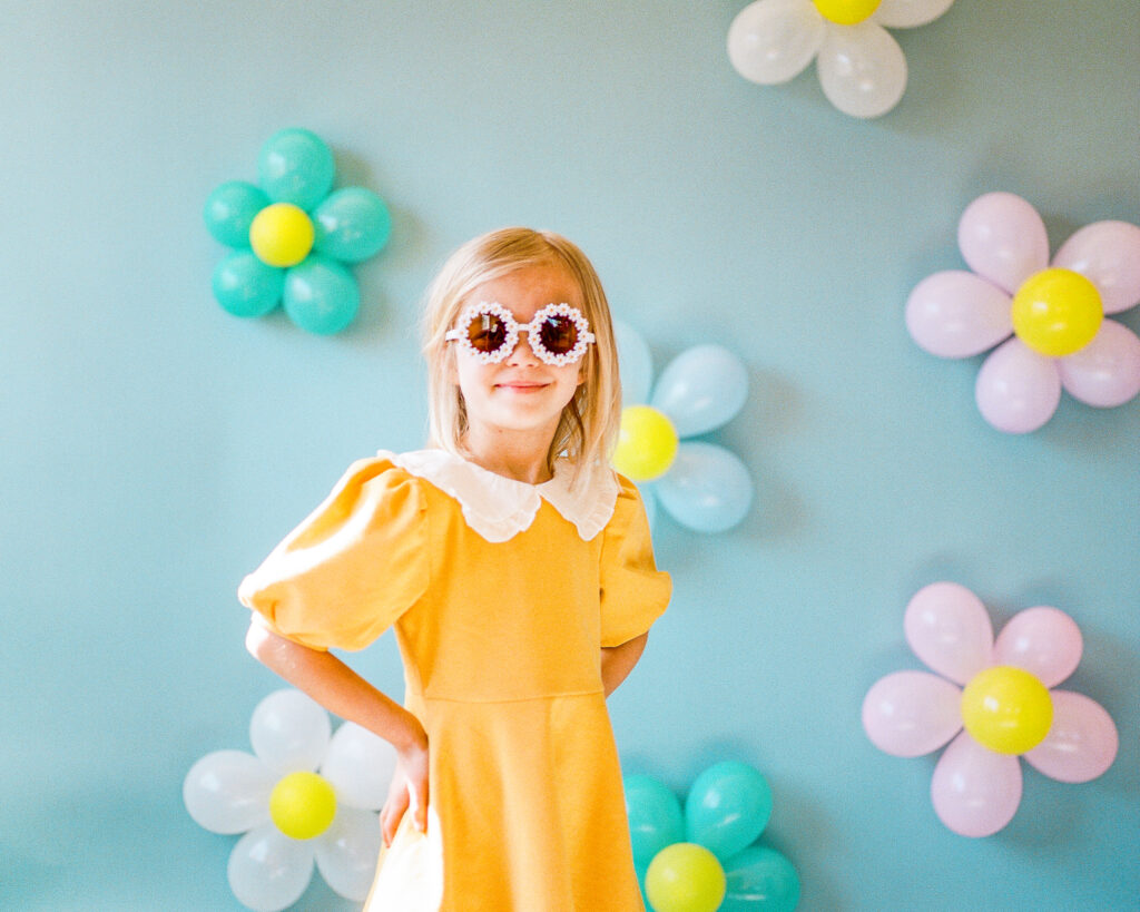 Family Photographer, a girl wears a yellow dress, sunglasses, and stands before a wall with balloons shaped as flowers